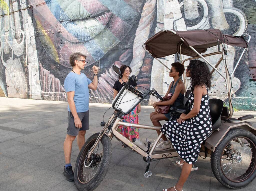 Four people in front of a painted mural depicting an Ethiopian woman's face, which is part of the Etnias Mural in Rio's Olympic Boulevard. A man in a blue t-shirt is standing in front of a pedicab talking to two women who are seated on the pedicab, and one who is standing beside it. The man is wearing a blue t-shirt and the most prominent of the women is wearing a navy blue dress with white polka dots.